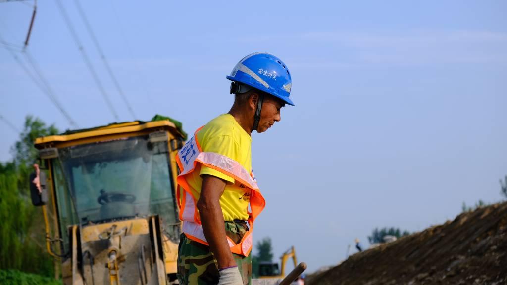 图片拍摄于江苏省宿迁市沭阳县京沪高速公路改扩建工程施工现场，一名建筑工人正在烈日下勤奋工作，皮肤被晒得黝黑，制度马甲从肩膀滑落。