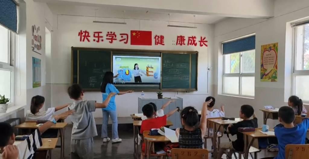 图为志愿者教小朋友们学习英语字母。志愿者用抽象的姿势让小朋友们认识字母，小朋友们积极与老师进行互动。